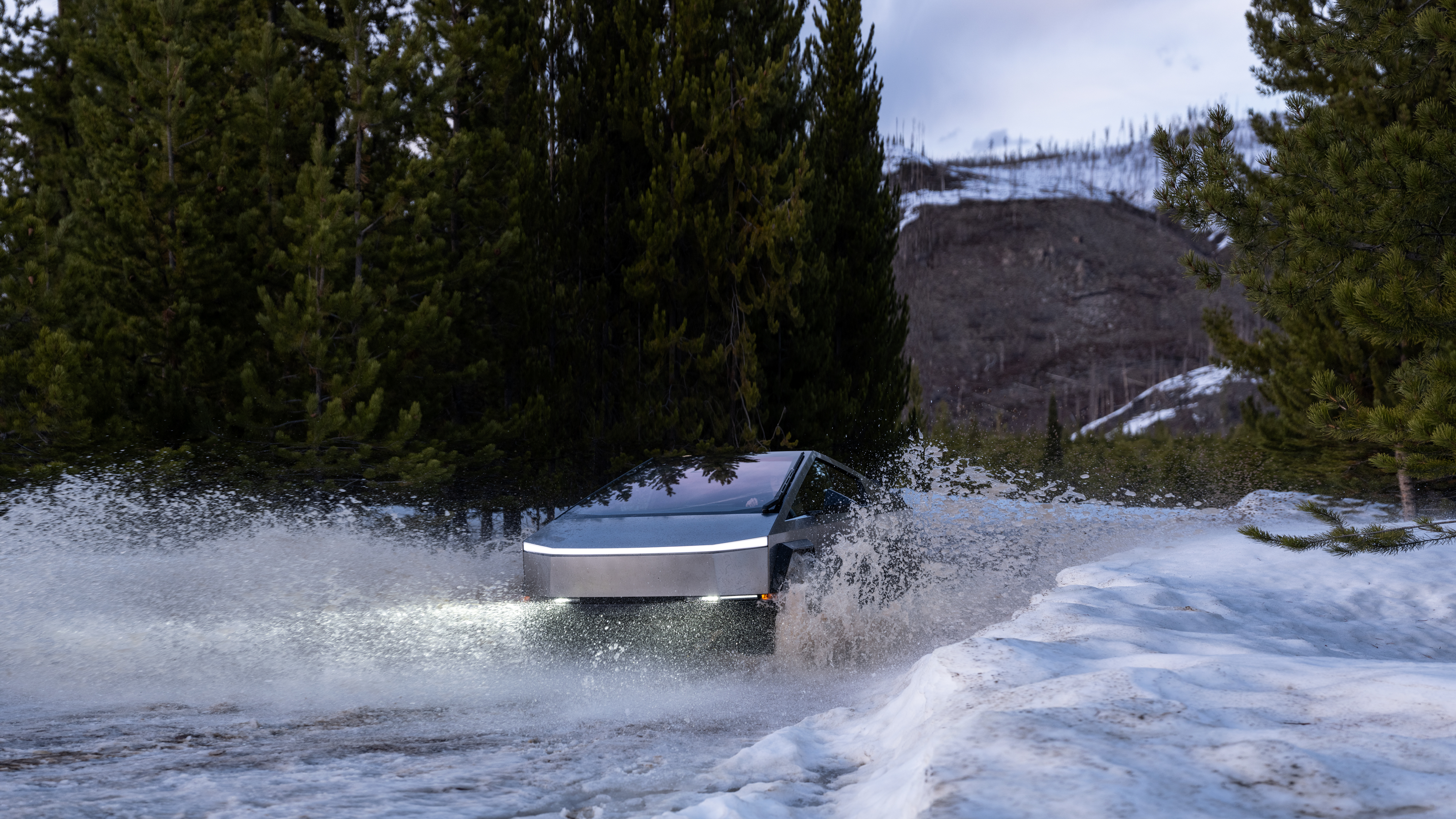 Cybertruck kör på en snöig och vattnig väg med grönskande träd i bakgrunden.