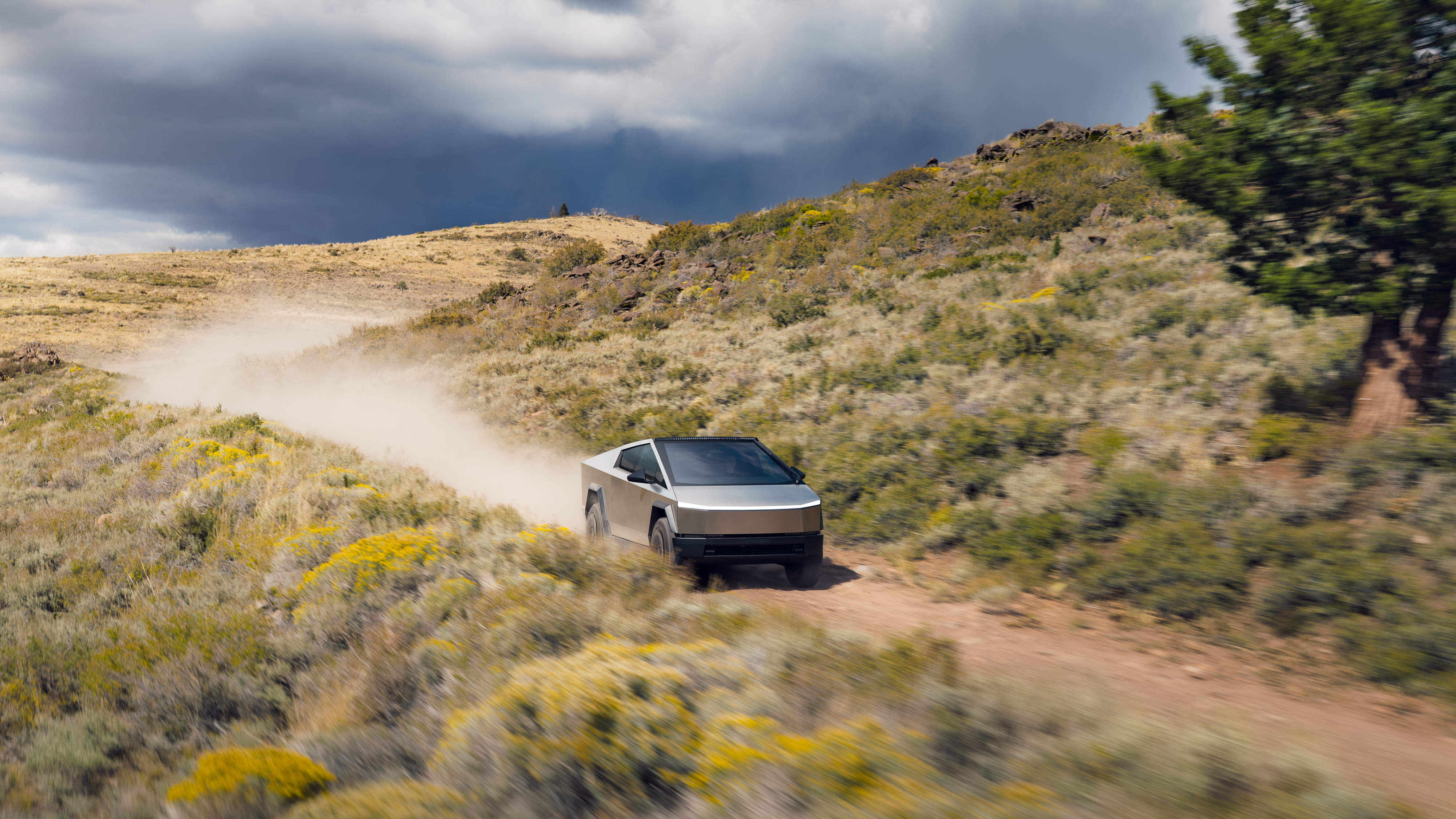 En vinklad bild av en Tesla Cybertruck som kör på en grusväg, med dammvirvlar och bergig terräng under en molnig himmel.