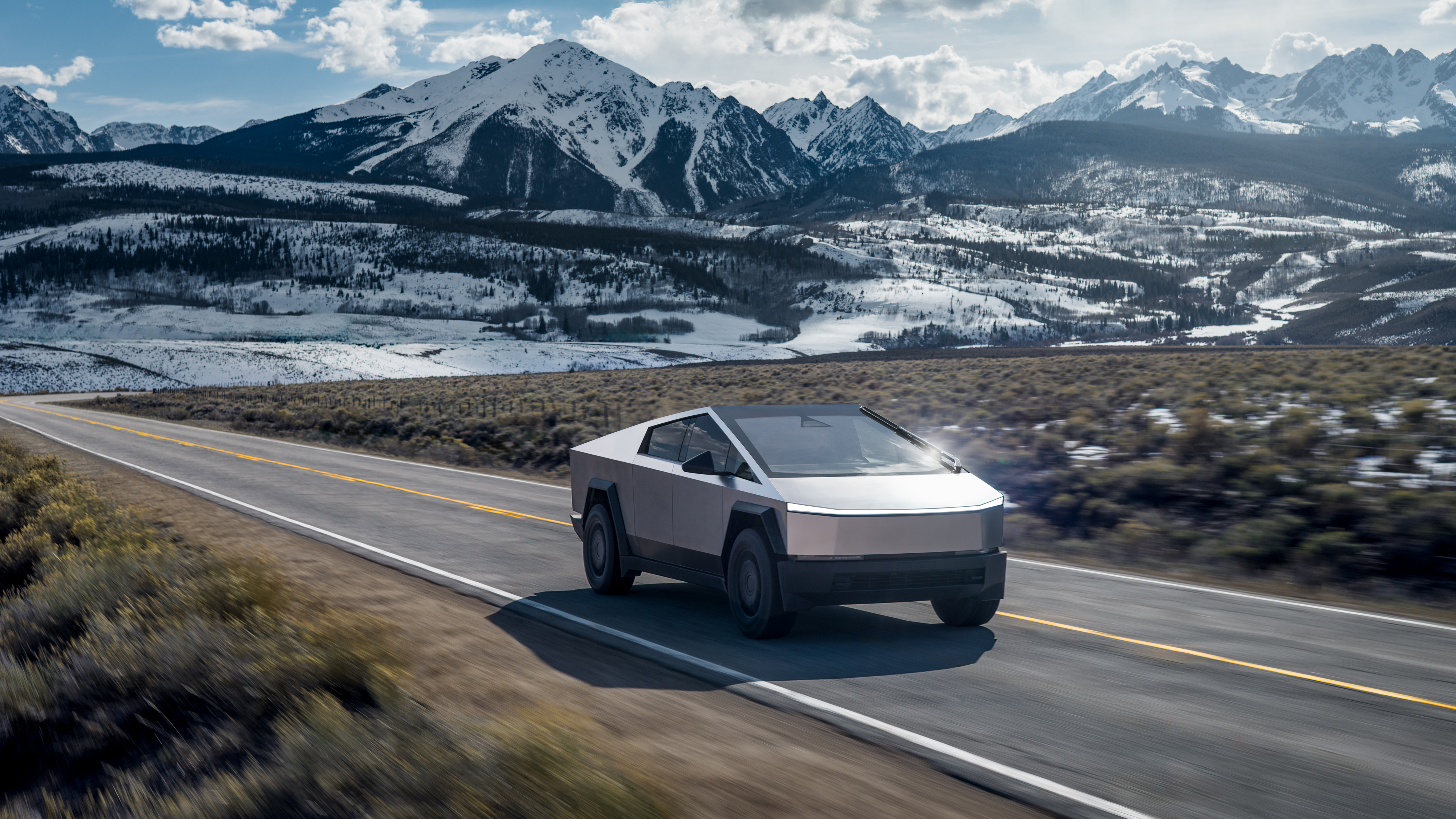 En Tesla Cybertruck körs på en asfalterad bergsväg med snötäckta berg i bakgrunden under en molnig himmel.