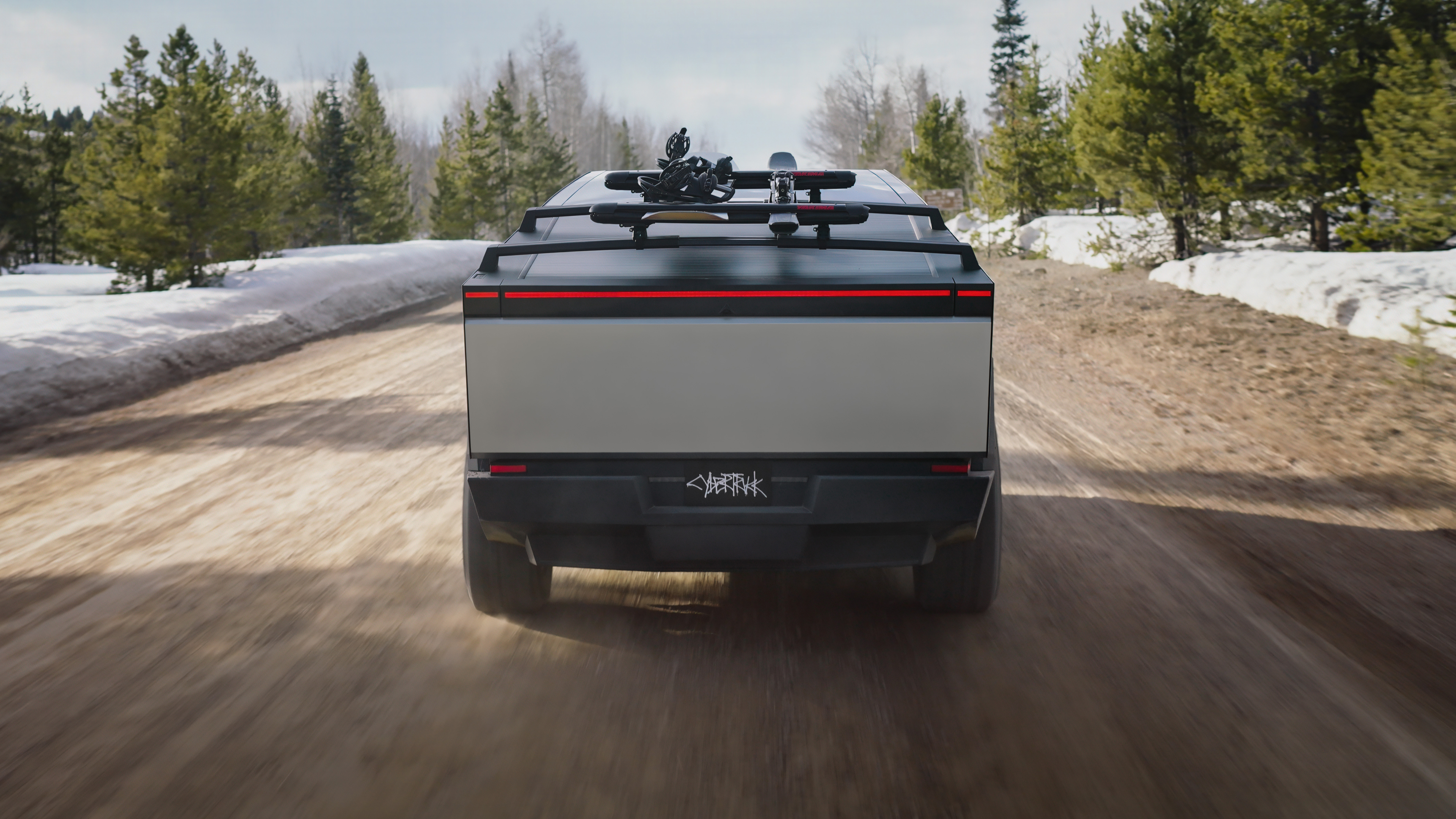 Tesla Cybertruck bakifrån med takräcke för snowboard och en lång röd ljuslinje som bakljus i en skogsstig.