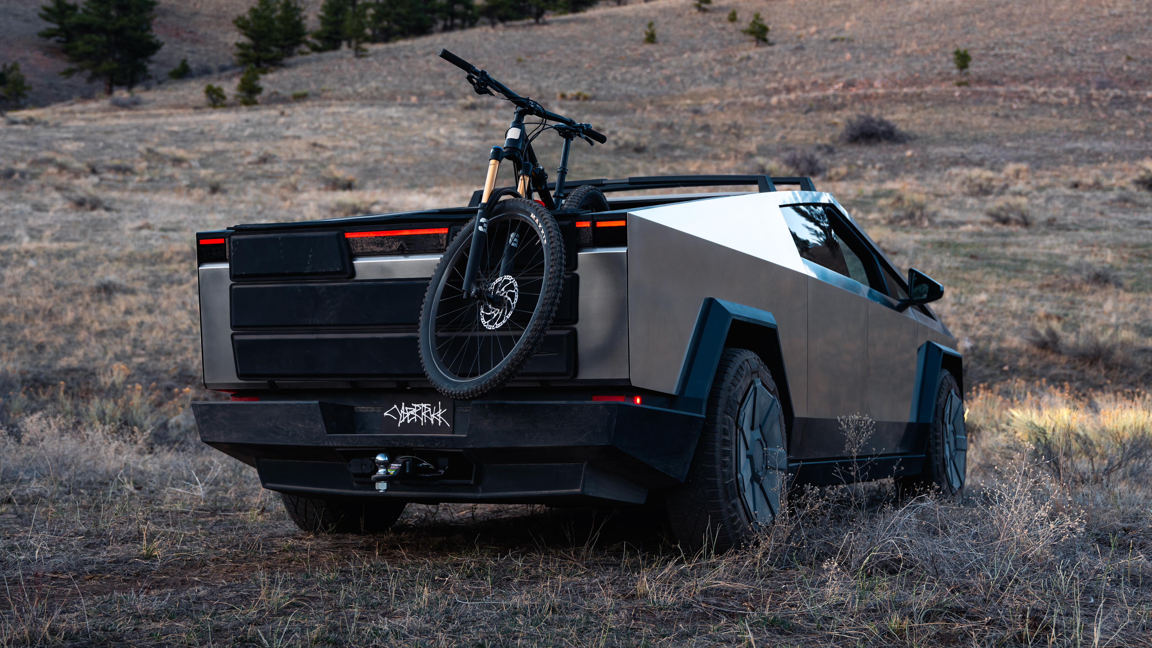 En monterad mountainbike på lastluckan av en grå Tesla Cybertruck med bakljusen tända, på ojämn mark.