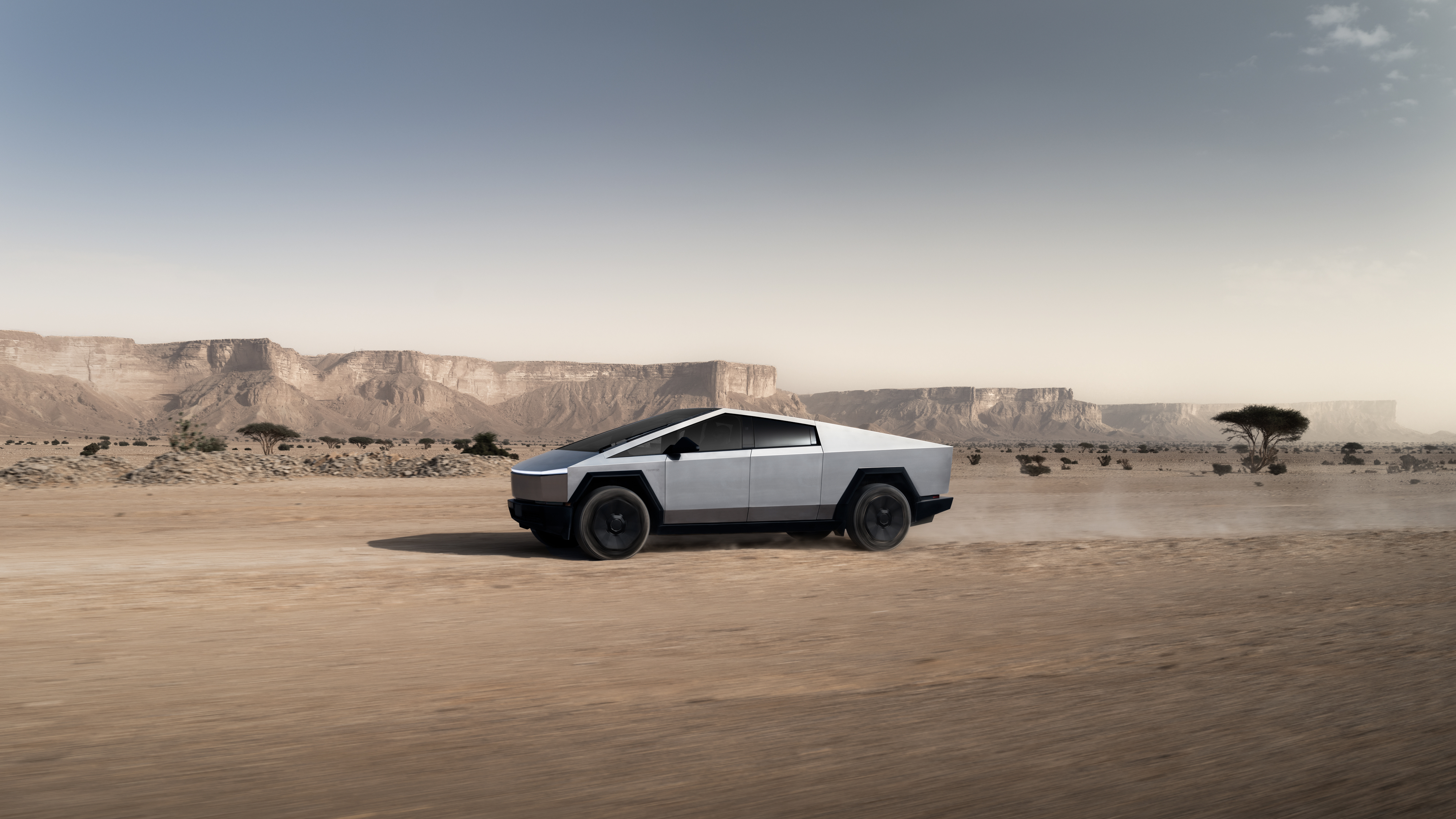 En Tesla Cybertruck ses från sidan som kör på en grusväg framför berg i ett kargt ökenlandskap under en molnig himmel.