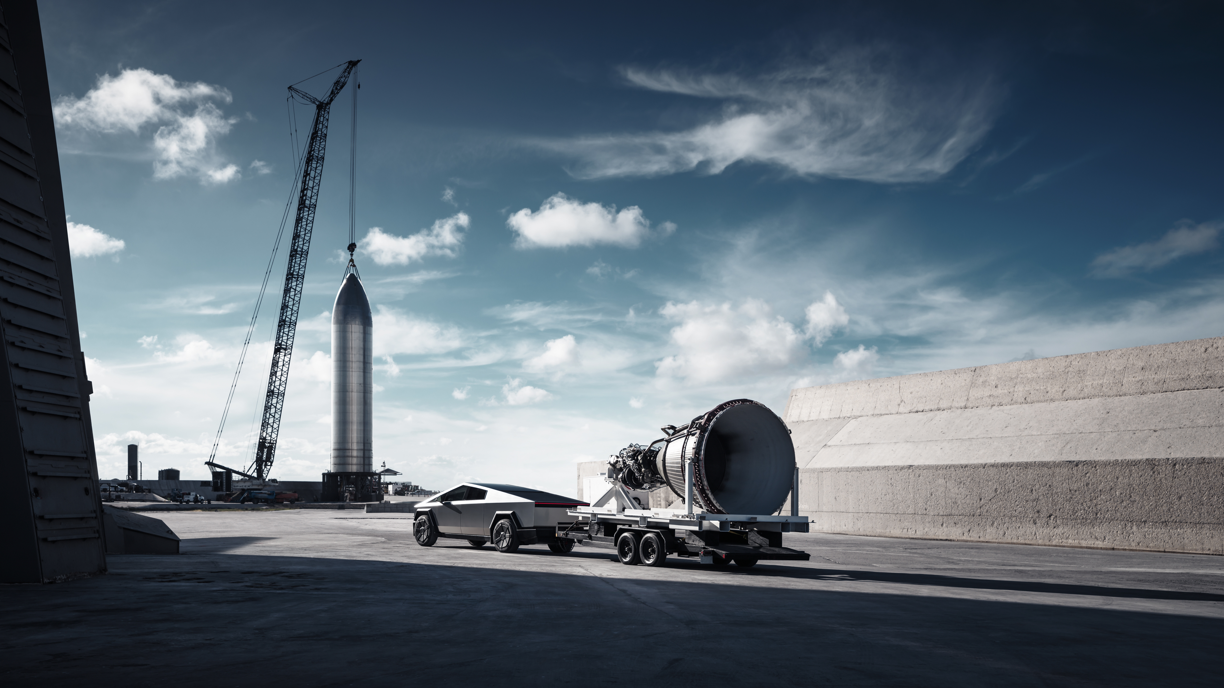 En silverfärgad Tesla Cybertruck drar en stor raketmotor på en trailer under en blå himmel med tunna moln.