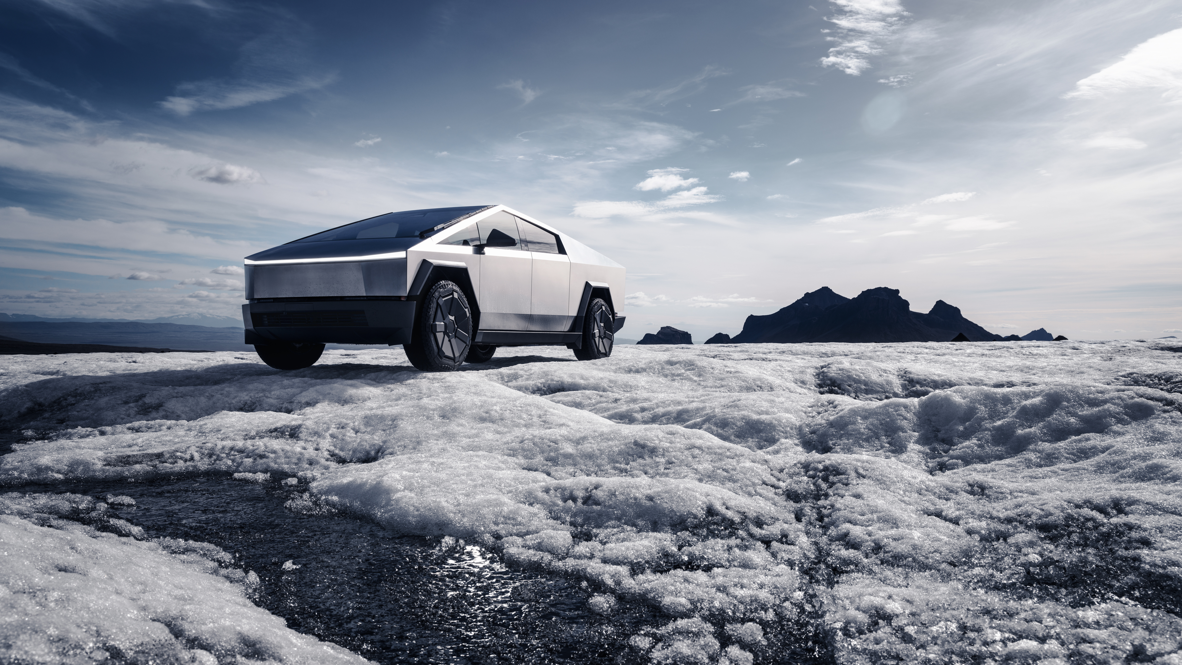 Tesla Cybertruck på en isig yta med vattensamlingar i förgrunden och bergiga formationer i bakgrunden under en molnig himmel.