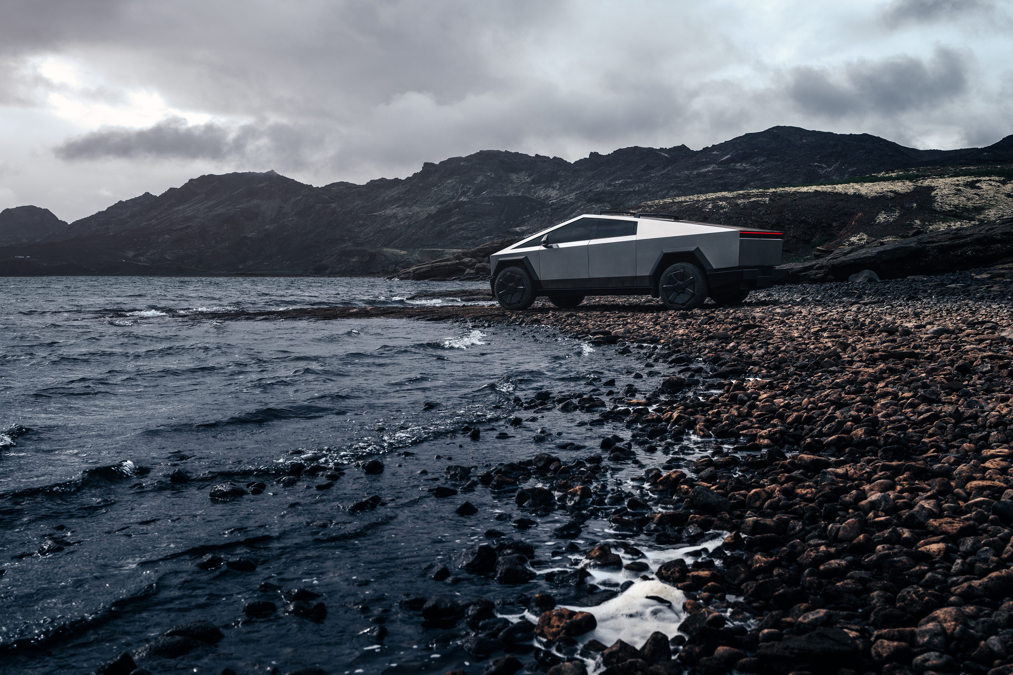 En Cybertruck står parkerad på en stenig strand, med havet och mörka berg i bakgrunden under en mulen himmel.