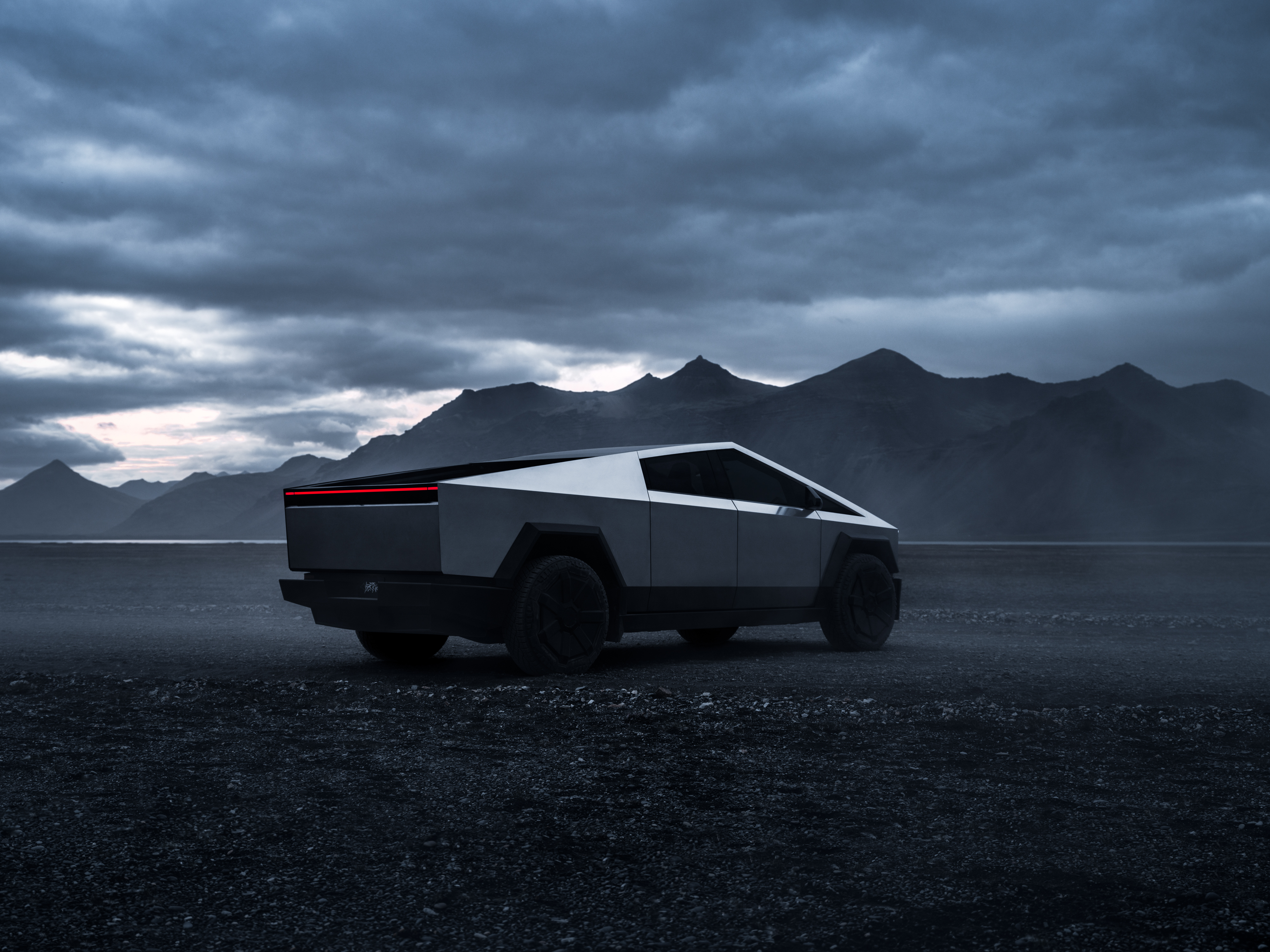 En Tesla Cybertruck står i profil på en grusväg, med berg och molnig himmel i bakgrunden.