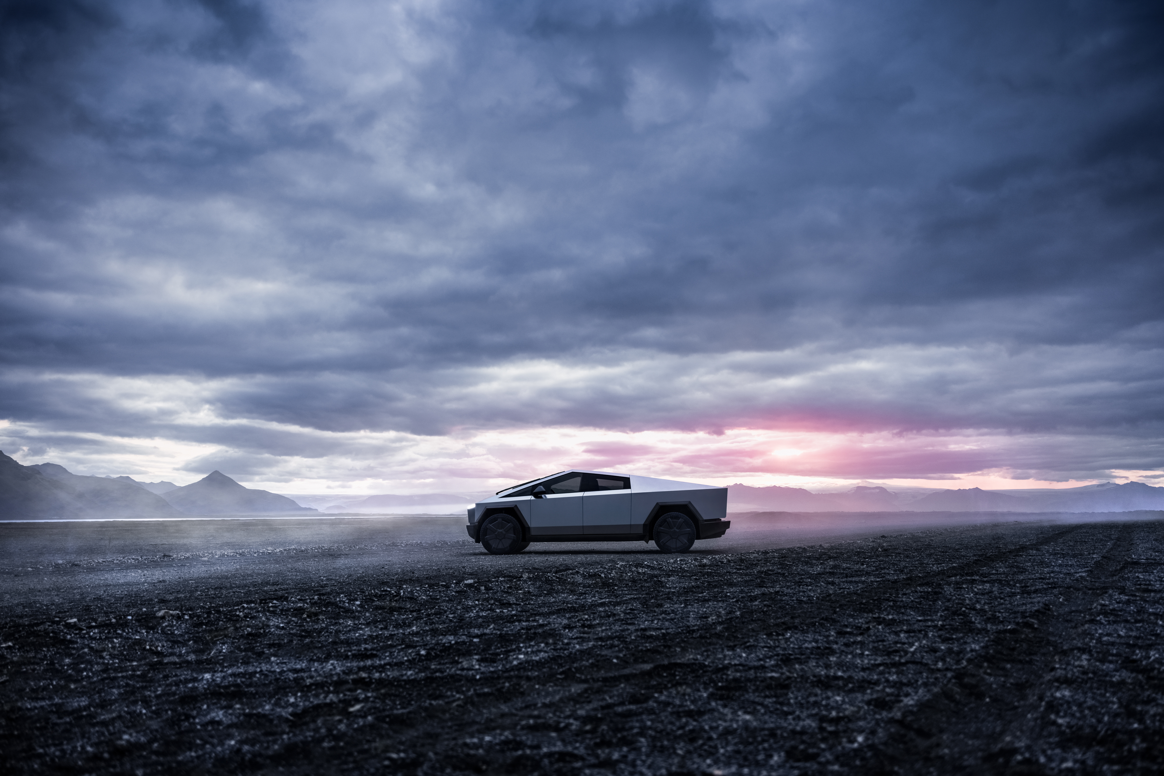 En sidovy av en Tesla Cybertruck stående på stenig mark under en dramatisk, molnig himmel med inslag av rosa.