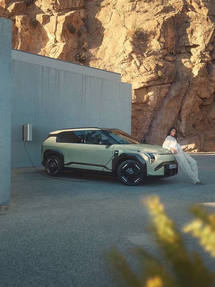 Kia EV3 ser ut att bli en prisvärd elbil med attraktiv design. Här fotograferas den under laddning, med en kvinna poserande mot bilens front.